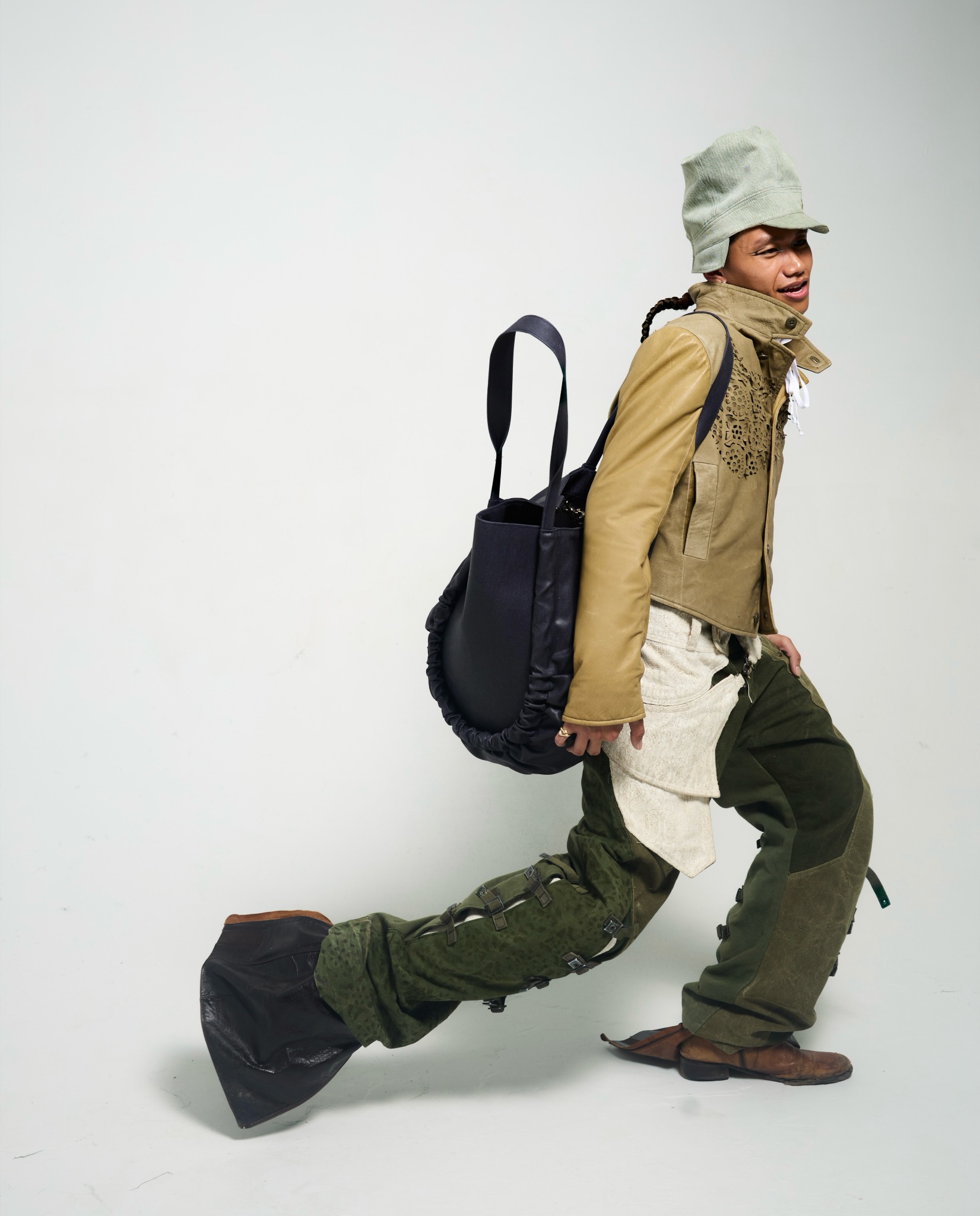 OSČ lookbook: green jacket and buckled trousers with an oversized dark shoulder bag, photographed in motion.